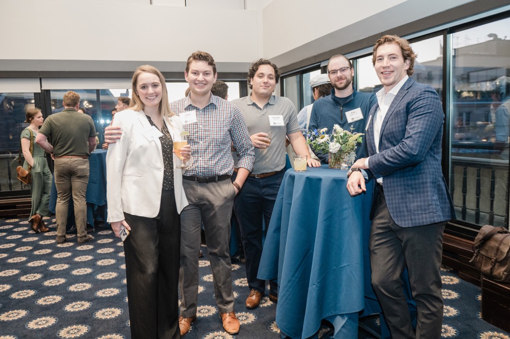 GVSU Alumni stand smile at camera and stand around table. One man is leaning on table.